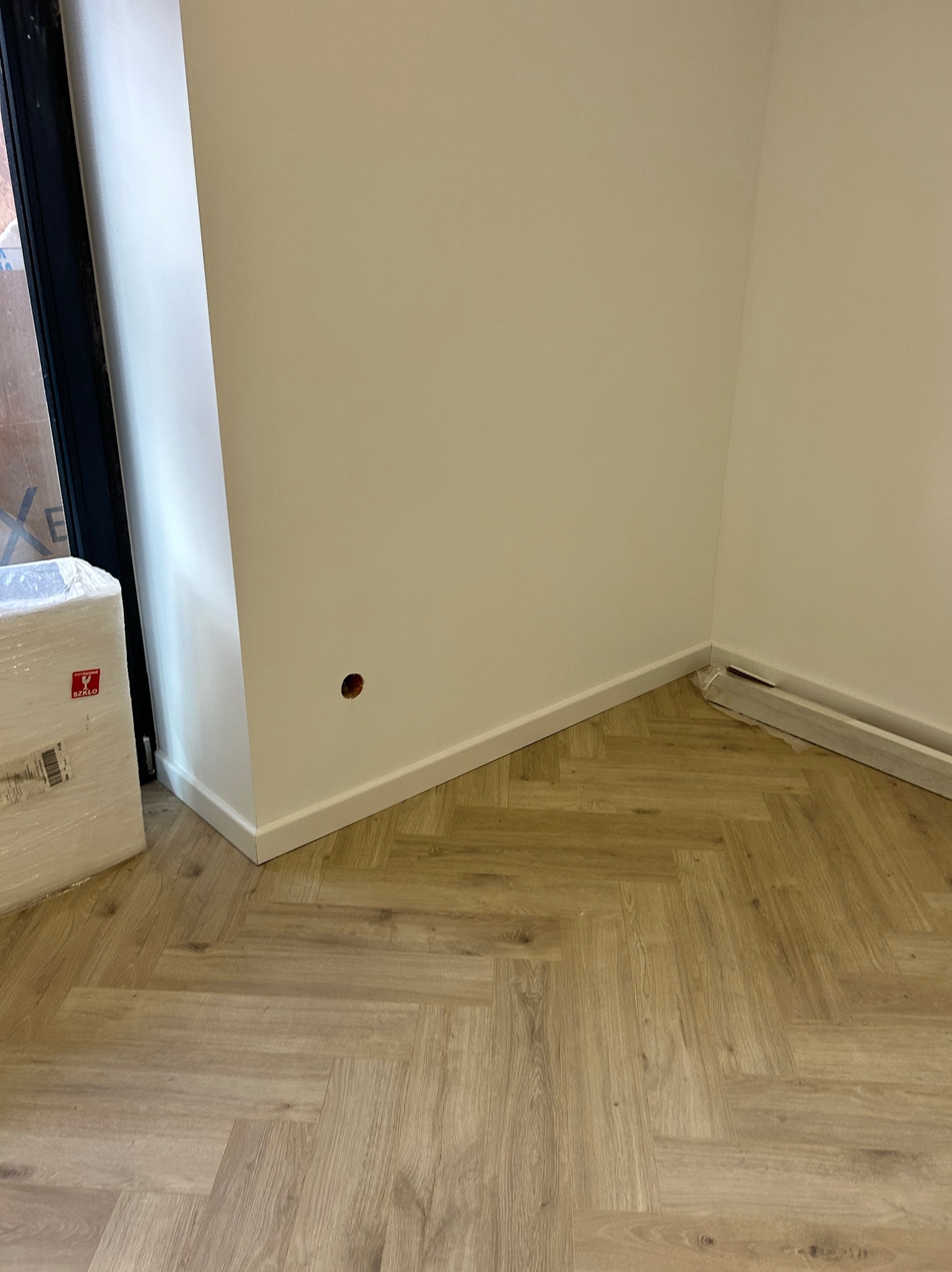 Pomalowane na biało ściany w rogu pomieszczenia z nową podłogą ułożoną w jodełkę; widoczna puszka z elektryką i zapakowane elementy przypodłogowe.