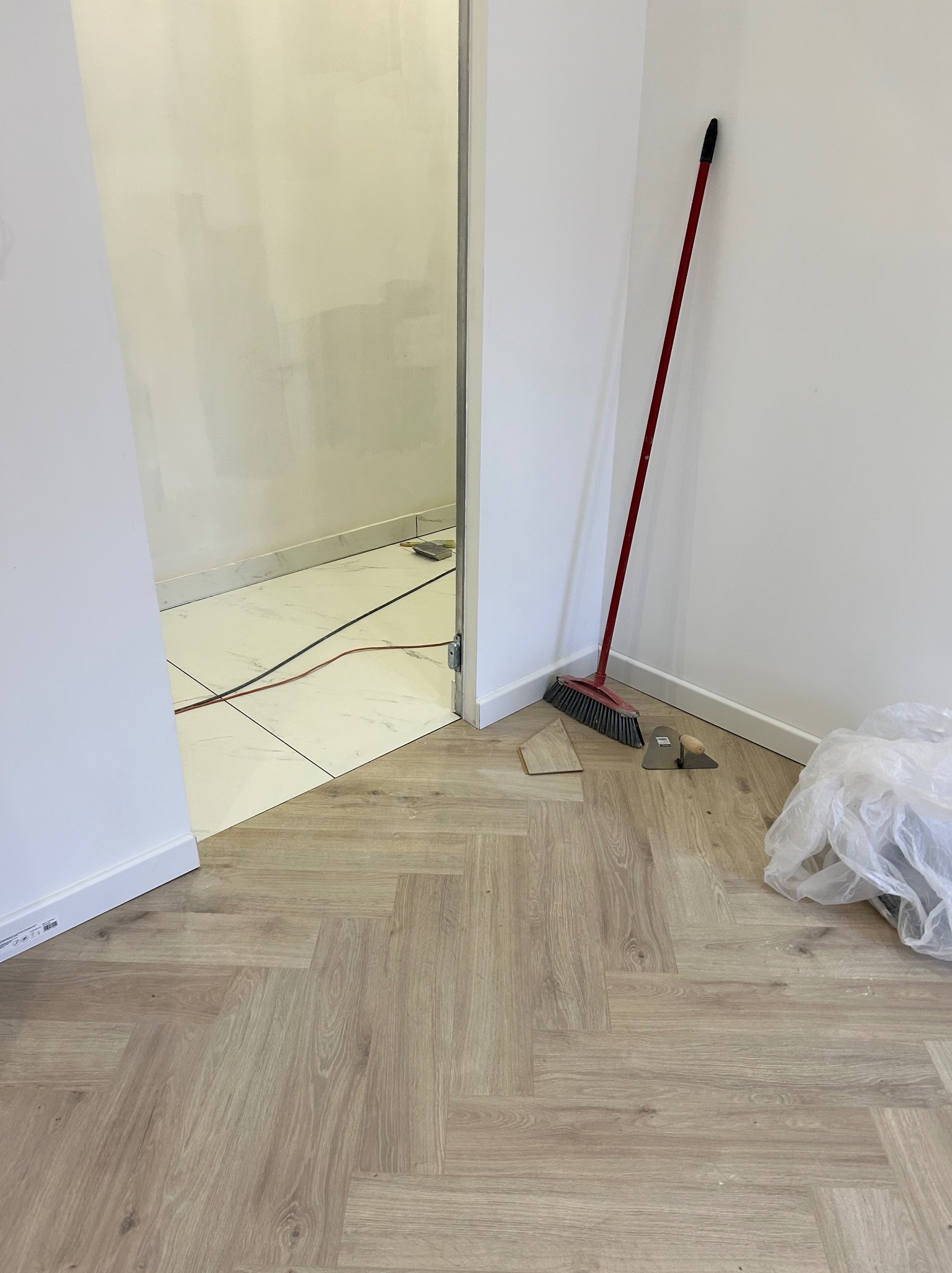 Podłoga z parkietu ułożonego w jodełkę, przejście do łazienki z białymi płytkami, czerwona zmiotka oparta o ścianę, kielnia i fragment płytki na podłodze, worek z odpadami po remoncie.