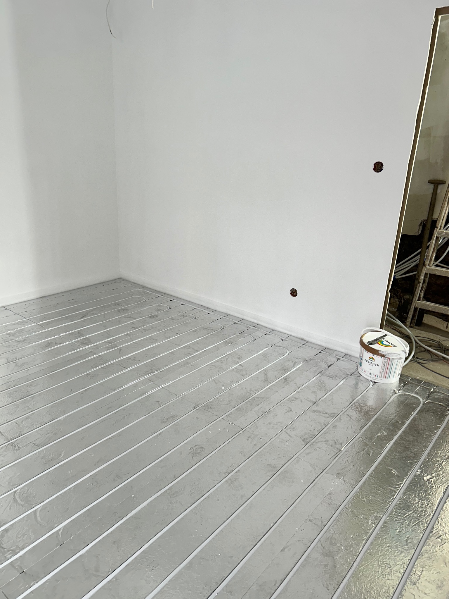 Przygotowanie do ogrzewania podłogowego: srebrna folia izolacyjna z rurkami ułożona na podłodze w rogu pokoju, obok puszka z białą farbą Designer White.