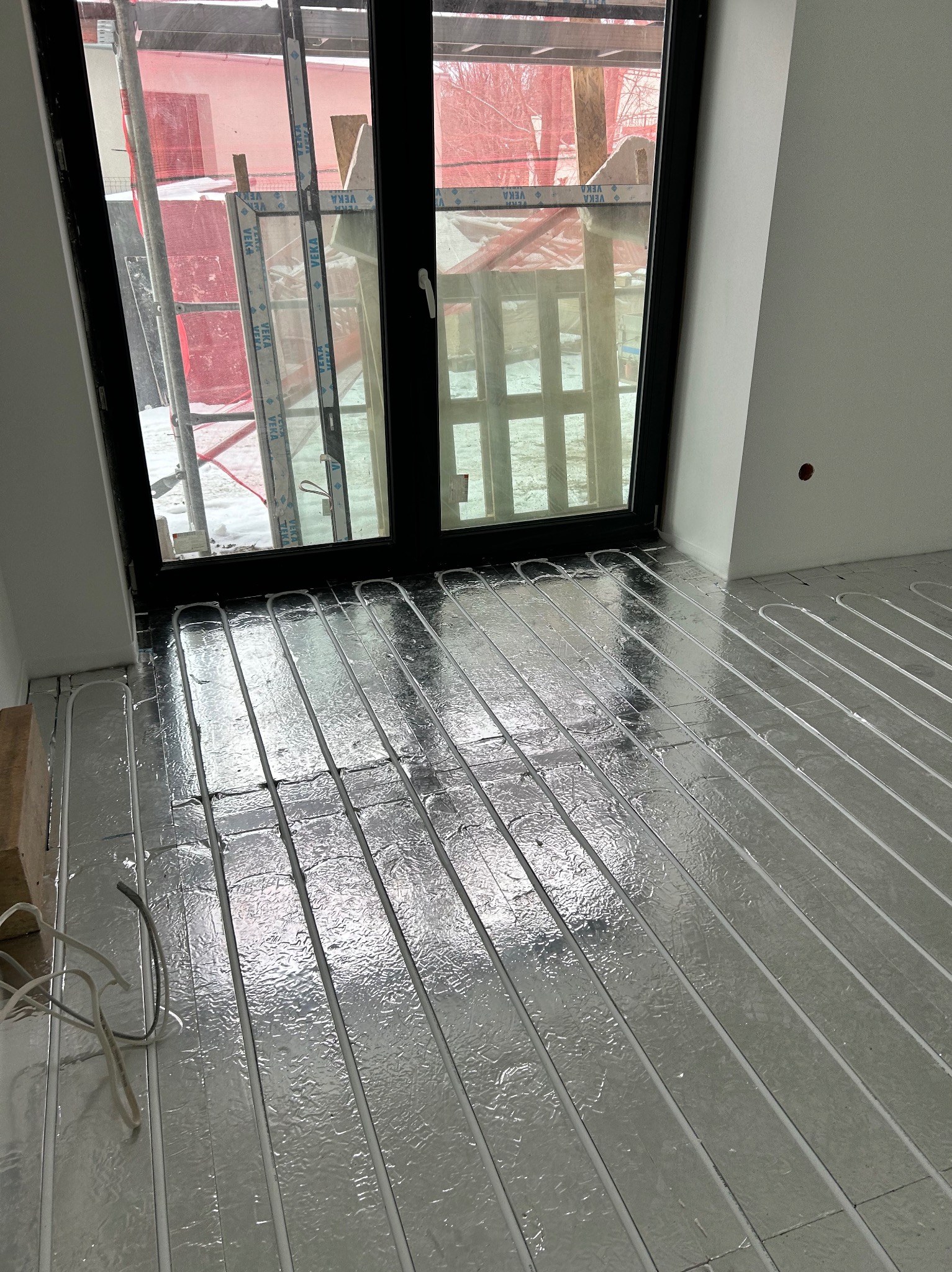 Instalacja ogrzewania podłogowego: białe rurki układane na srebrnej folii izolacyjnej w pomieszczeniu z dużym oknem, widok na zewnątrz z rusztowaniem i śniegiem.