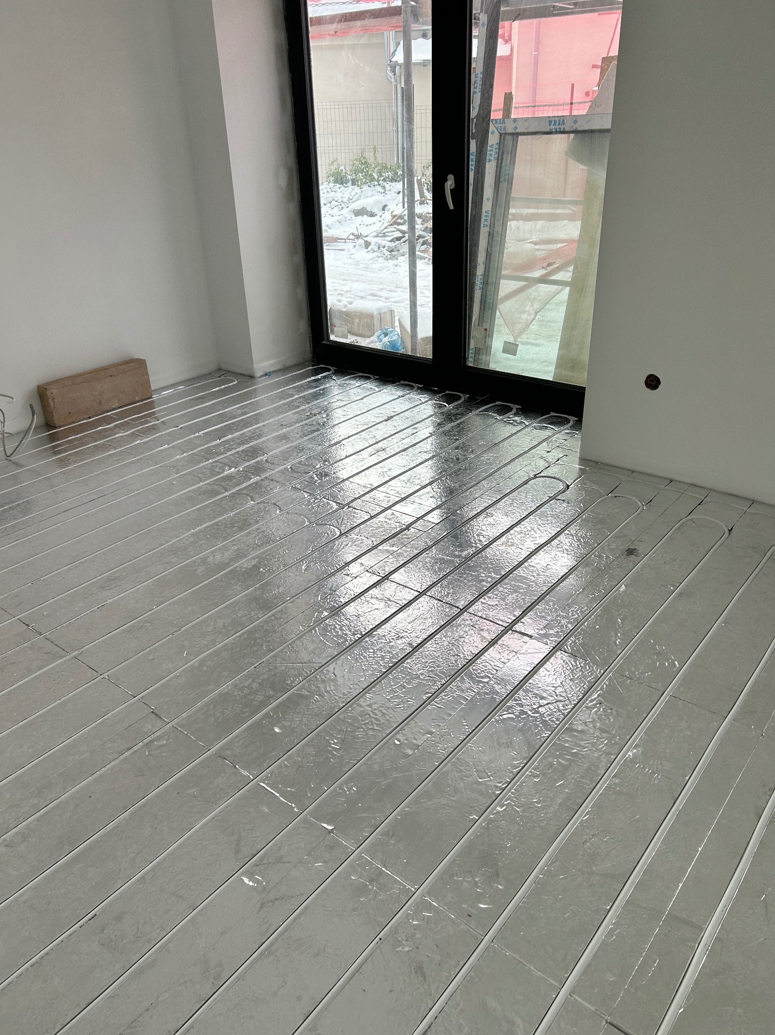 Montaż ogrzewania podłogowego w nowym pomieszczeniu, widoczne rury grzewcze ułożone na folii izolacyjnej, cegła oparta o ścianę, okno balkonowe z widokiem na podwórze.