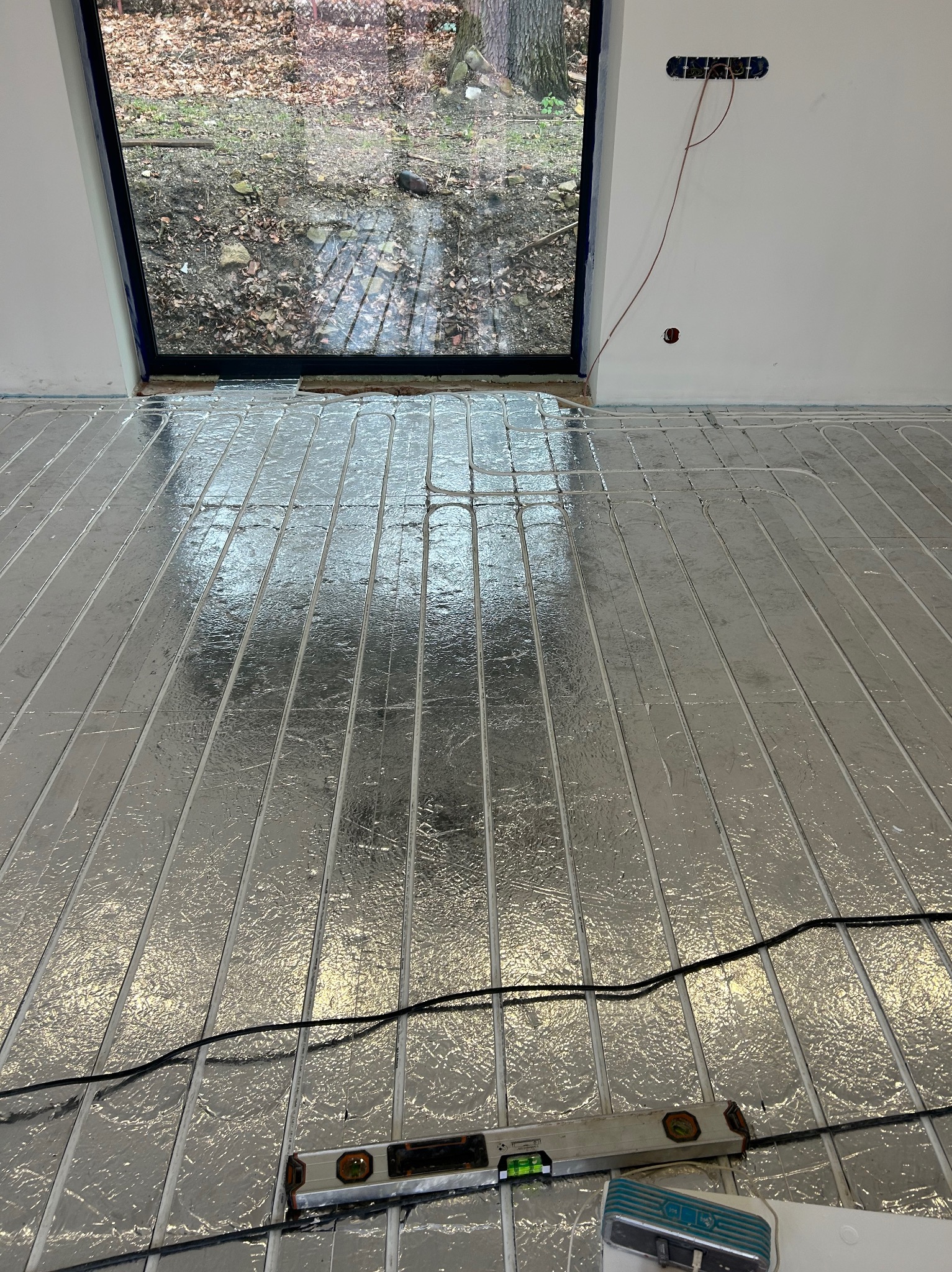 Instalacja folii grzewczej na podłodze z widocznym poziomem i przewodami elektrycznymi, okno z widokiem na ogród w tle.