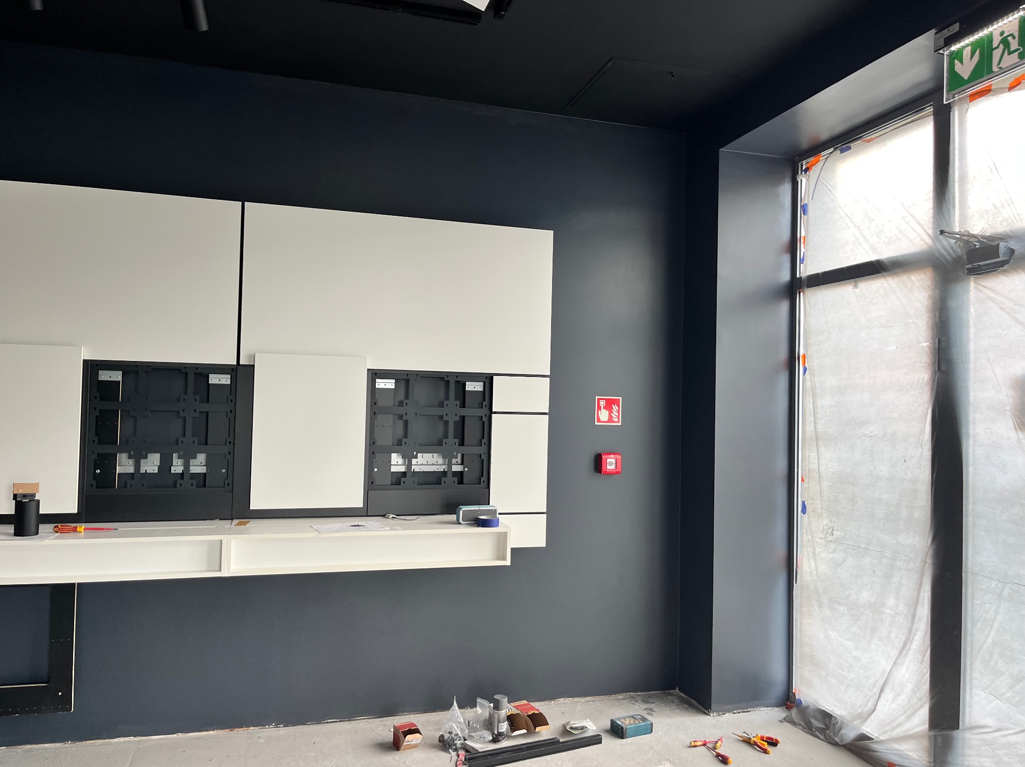 Montaż białych paneli ściennych z czarnymi wnękami na szarej ścianie biura, widoczne narzędzia i akcesoria na podłodze, okno zabezpieczone folią.