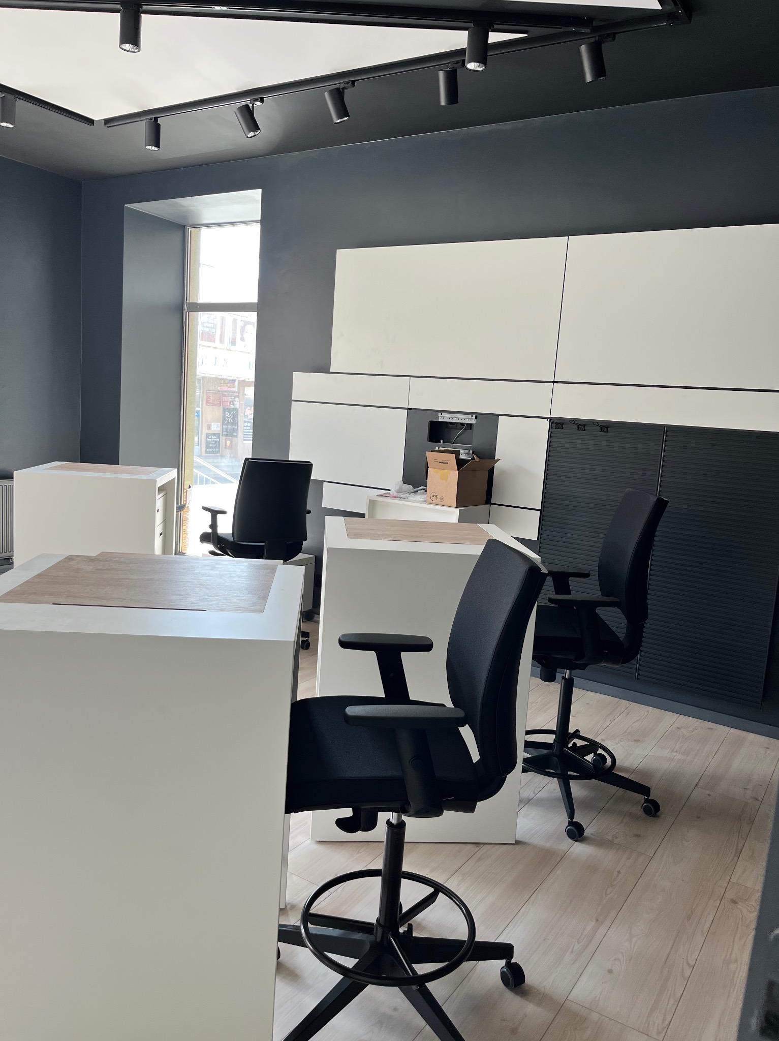 Wnętrze biura w trakcie urządzania: minimalistyczne białe biurka z drewnianymi blatami, czarne krzesła obrotowe, szare ściany i sufit z czarnymi reflektorami, białe szafki wiszące, karton na blacie.