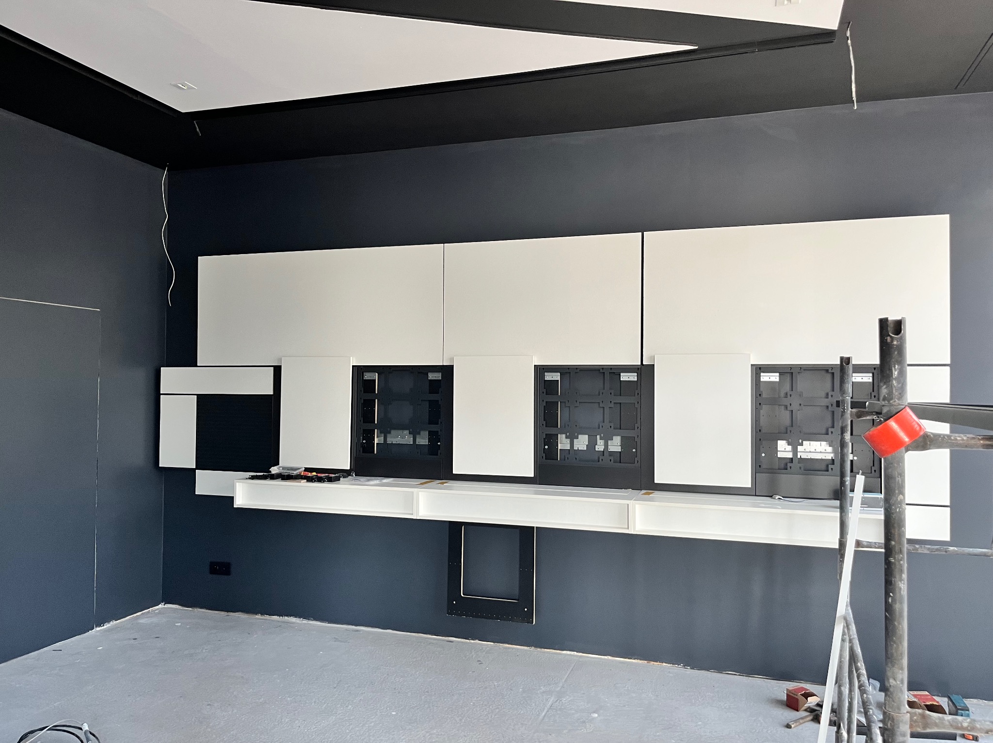 Montaż białych szafek wiszących na ciemnej ścianie, widoczne otwory na instalacje, rusztowanie z boku, surowy stan wykończenia podłogi.