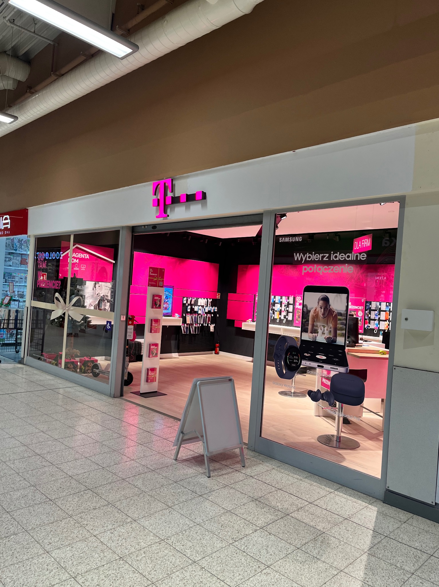 Wejście do salonu operatora telekomunikacyjnego T-Mobile z widocznym logo i wystawą smartfonów, zegarków i słuchawek wewnątrz, prezentowanych na tle intensywnego różowego oświetlenia.