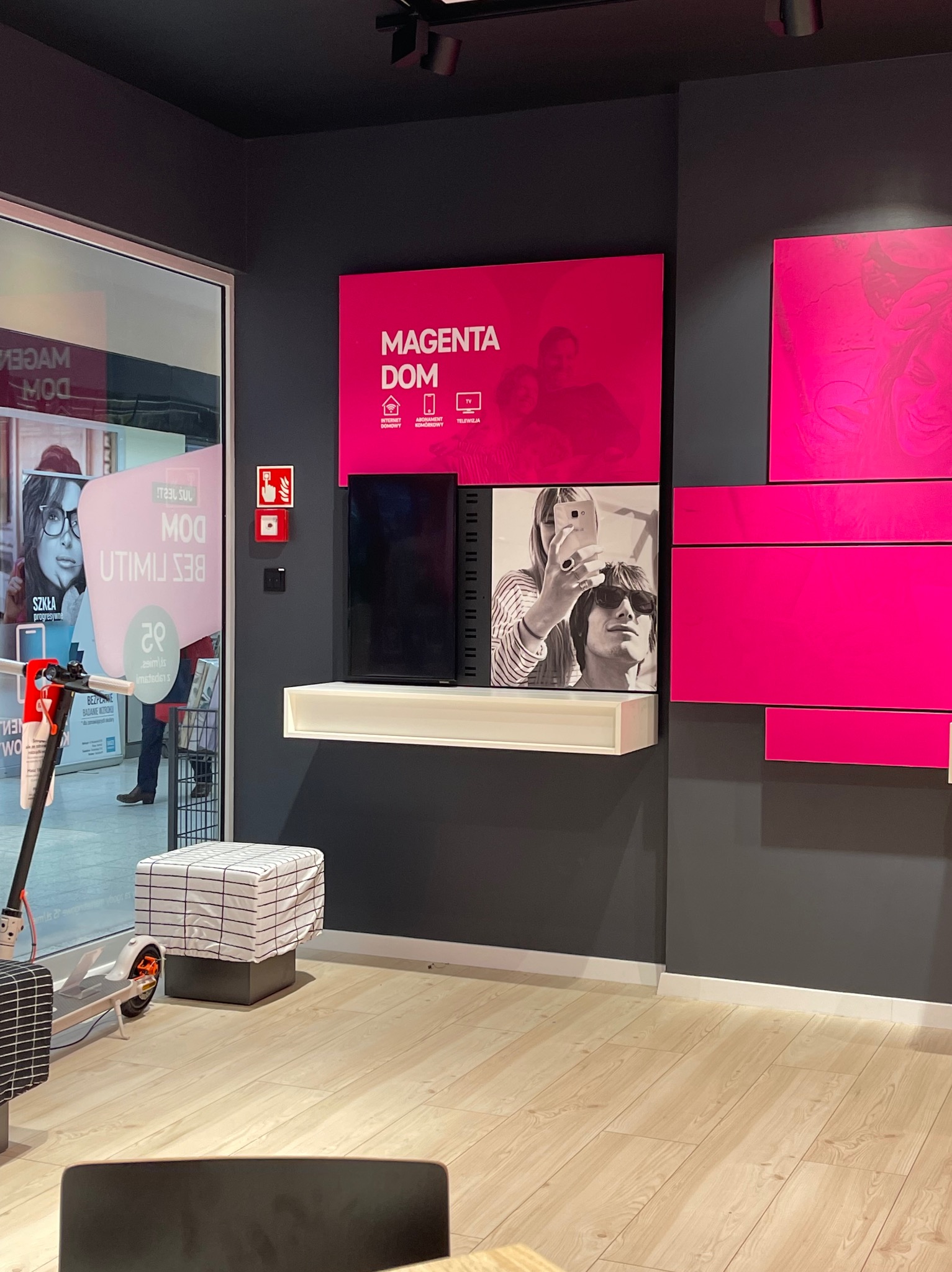 Wnętrze punktu obsługi klienta operatora telekomunikacyjnego z elementami identyfikacji wizualnej marki Magenta Dom, w tym plakaty i aranżacja przestrzeni.