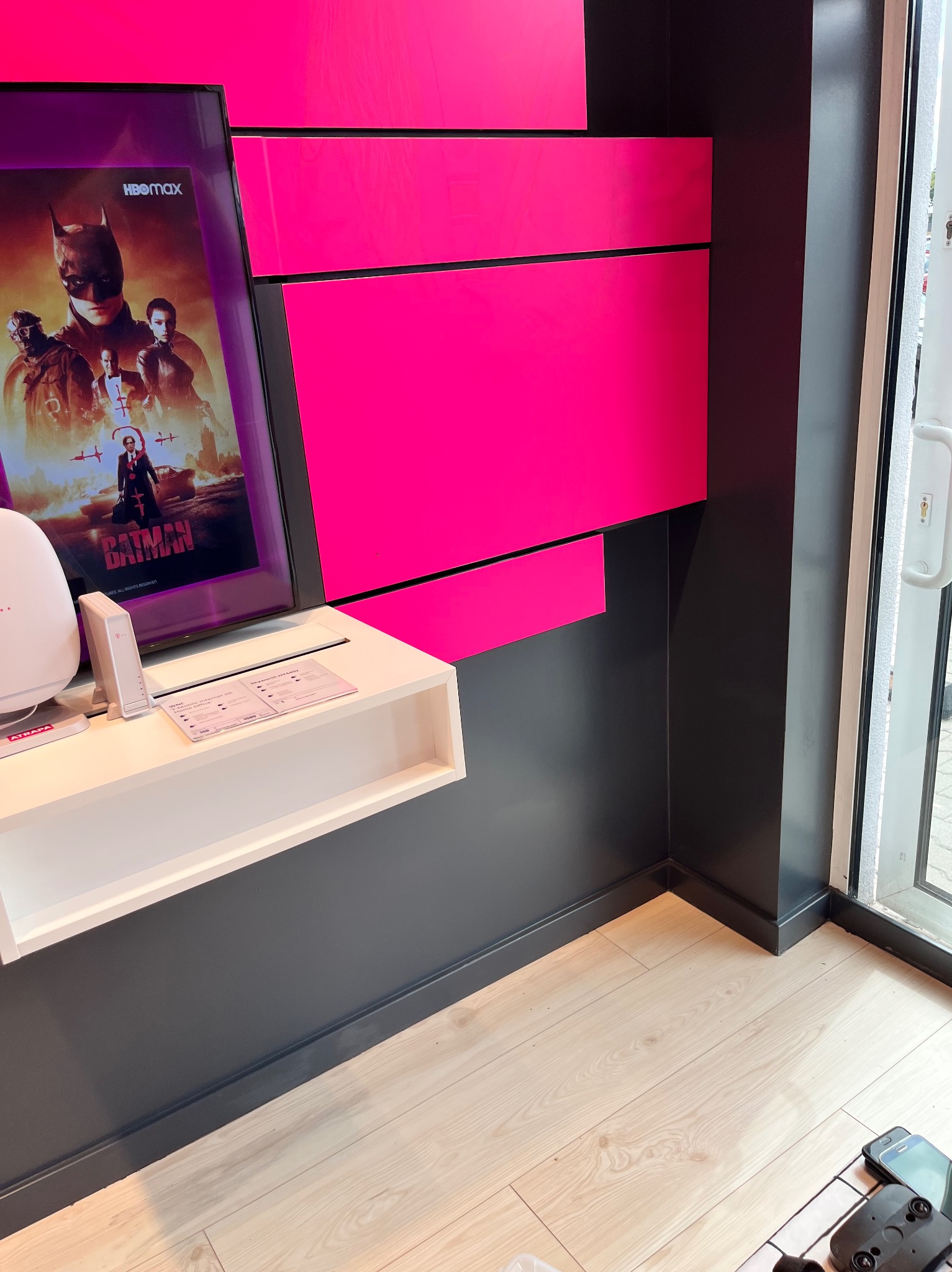 Wnętrze lokalu usługowego z elementami w kolorze fuksji: półka z routerem i ulotkami reklamowymi pod ekranem z plakatem filmu Batman, ściany w odcieniach szarości i podłoga z jasnych paneli.