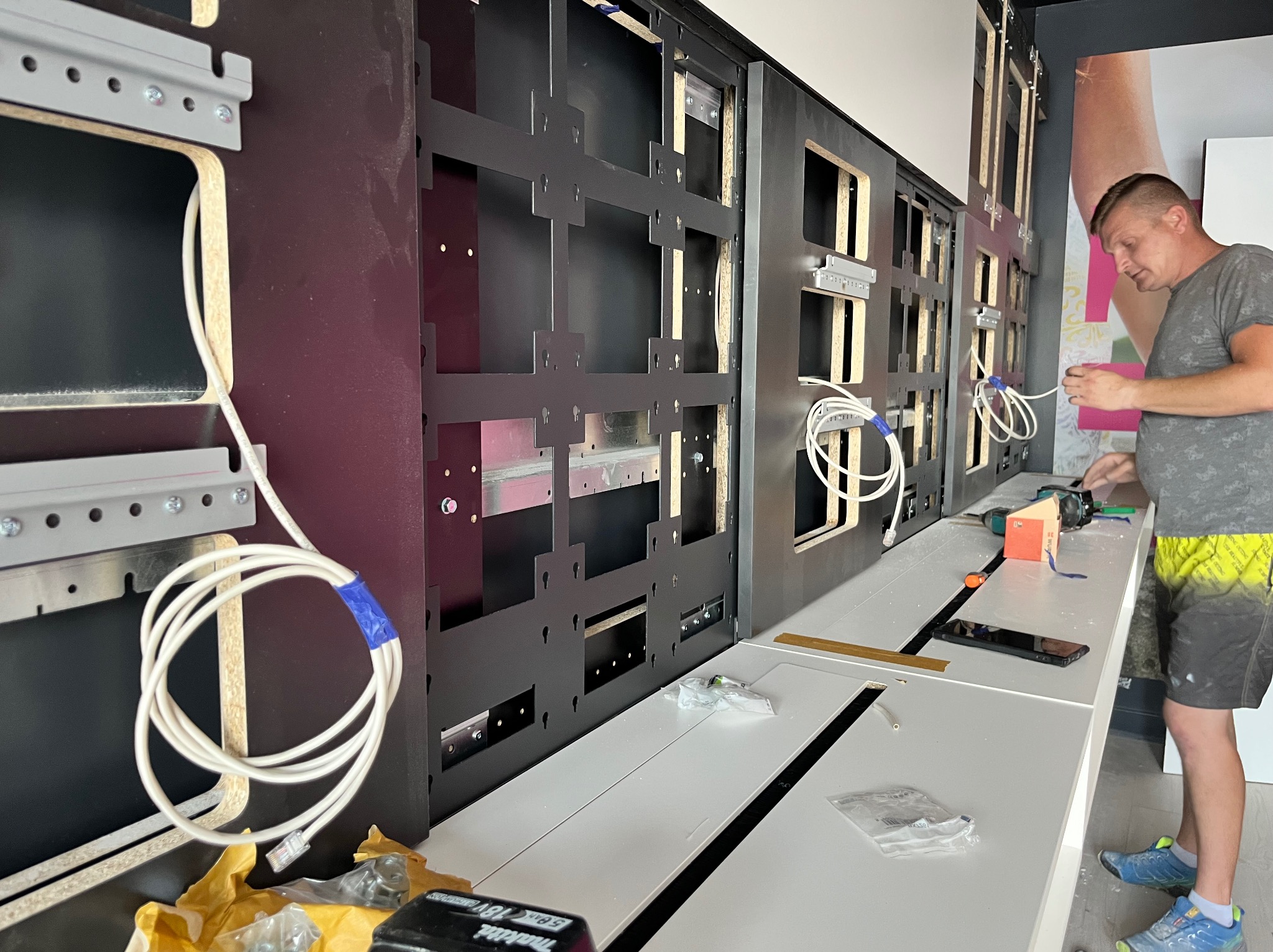 Mężczyzna instaluje metalową konstrukcję na ścianie z otworami, widoczne kable i narzędzia, etap montażu paneli ściennych w pomieszczeniu biurowym.