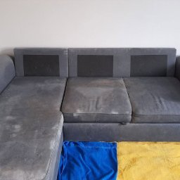 Mr. Glancer - Szara, narożna kanapa z widocznymi plamami przed praniem, zabezpieczona niebieskim i żółtym materiałem na podłodze.