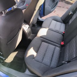 Mr. Glancer - Wnętrze samochodu z widocznymi tylnymi siedzeniami z ciemną tapicerką w odcieniach szarości i czerni, z rozłożonym podłokietnikiem środkowym i widocznymi czerwonymi zatrzaskami pasów bezpieczeństwa.