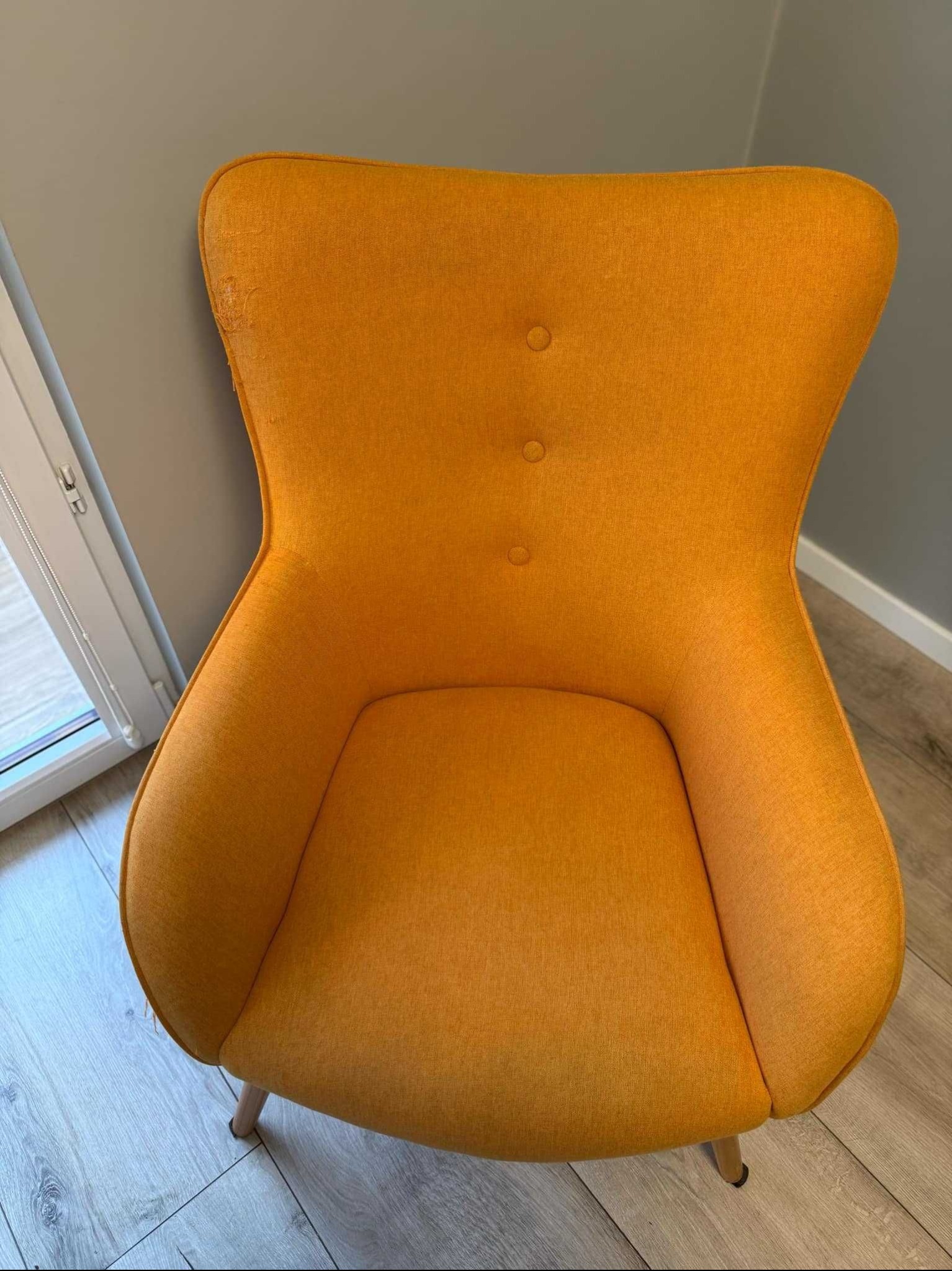 Żółty fotel tapicerowany z trzema guzikami na oparciu, widoczne uszkodzenie tapicerki na krawędzi oparcia, stojący na panelach podłogowych.