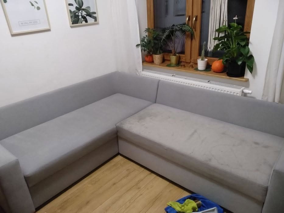 Szara narożna kanapa z widocznym, jaśniejszym, miejscowym zabrudzeniem na siedzisku, w tle okno z roślinami doniczkowymi i dekoracjami ściennymi.