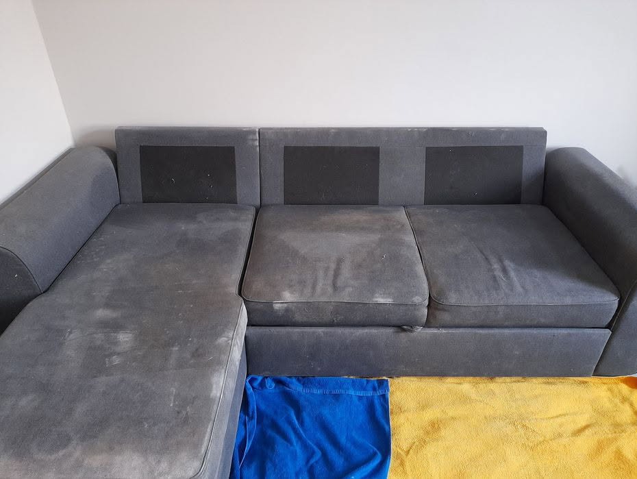 Szara, narożna kanapa z widocznymi plamami przed praniem, zabezpieczona niebieskim i żółtym materiałem na podłodze.