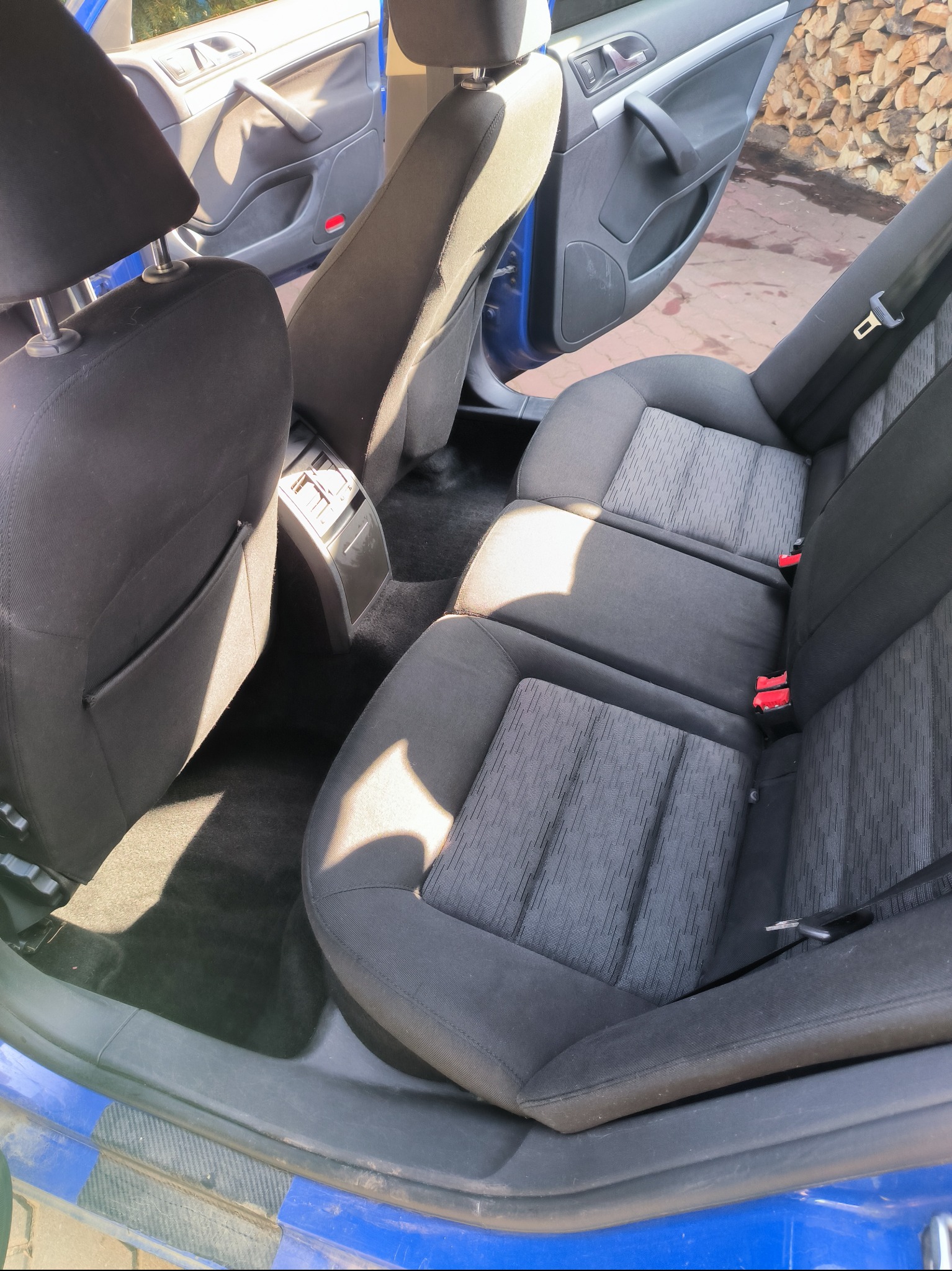 Wnętrze samochodu z widocznymi tylnymi siedzeniami z ciemną tapicerką w odcieniach szarości i czerni, z rozłożonym podłokietnikiem środkowym i widocznymi czerwonymi zatrzaskami pasów bezpieczeństwa.
