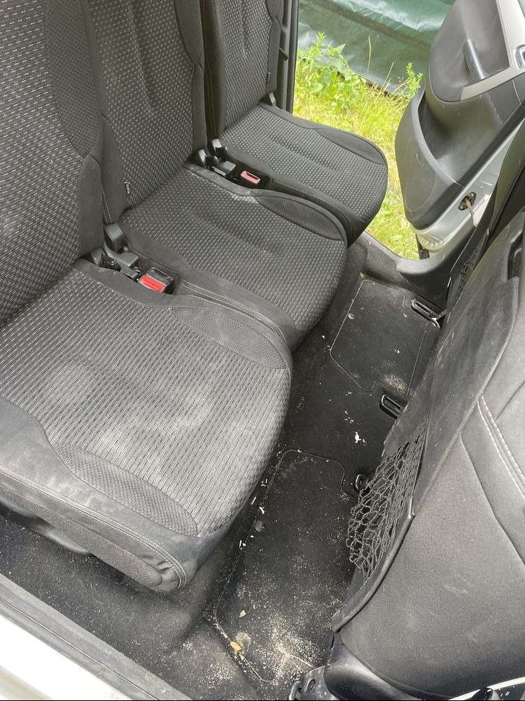 Wnętrze samochodu z widocznymi, czarnymi, tekstylnymi siedzeniami i zabrudzoną podłogą przed czyszczeniem. Widoczna siatka na drobiazgi po stronie pasażera.
