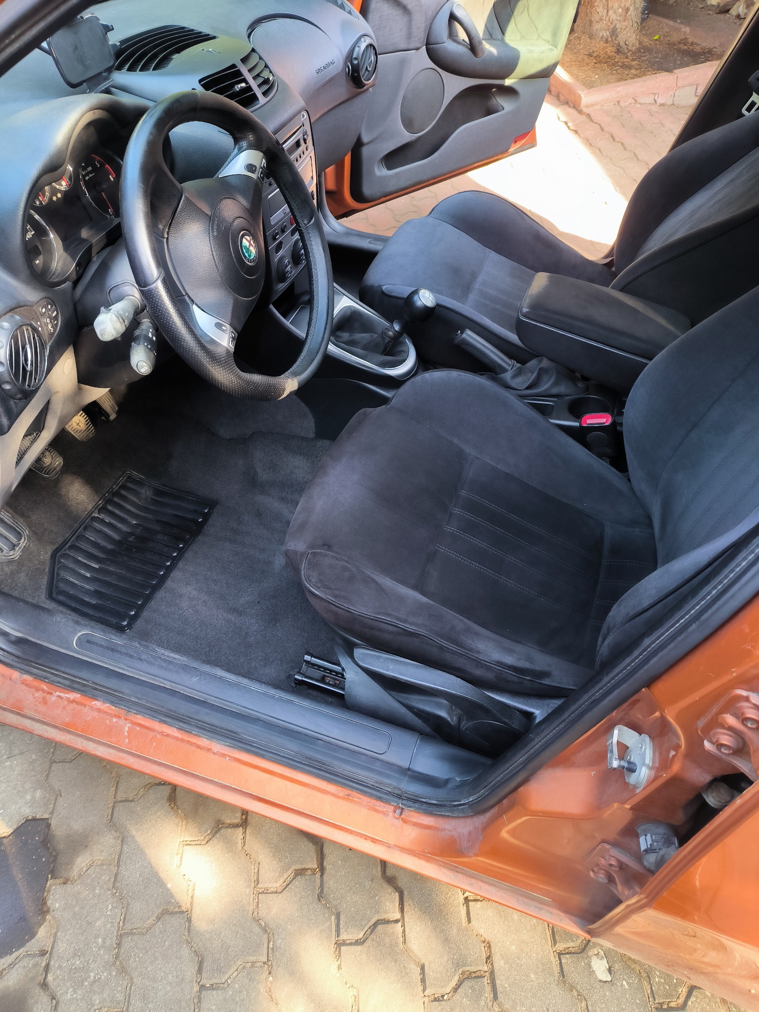 Wnętrze samochodu Alfa Romeo z czarną tapicerką welurową, widoczne fotele, dywaniki i deska rozdzielcza, pomarańczowy lakier karoserii.