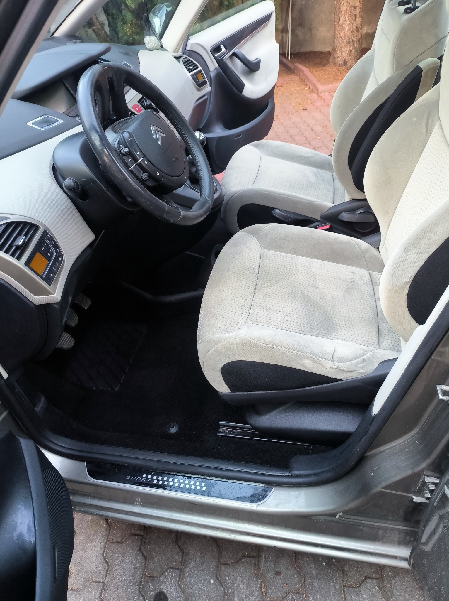 Wnętrze samochodu z beżowo-czarną tapicerką, widoczne fotele przednie, kierownica i deska rozdzielcza, otwarte drzwi od strony kierowcy.