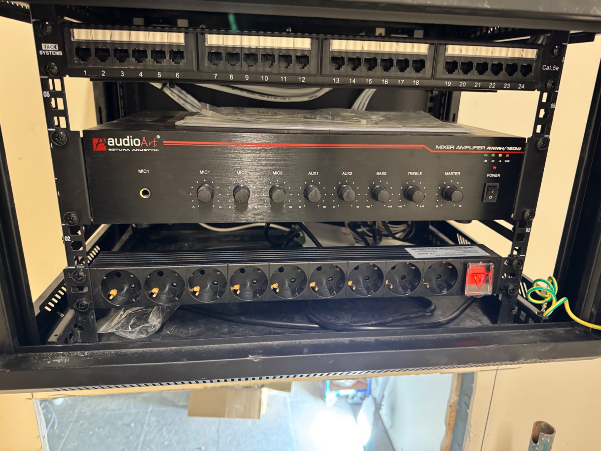 Panel krosowniczy, wzmacniacz audio i listwa zasilająca w szafie rack. Widoczne kable i złącza. Sprzęt audio zamontowany w metalowej obudowie.