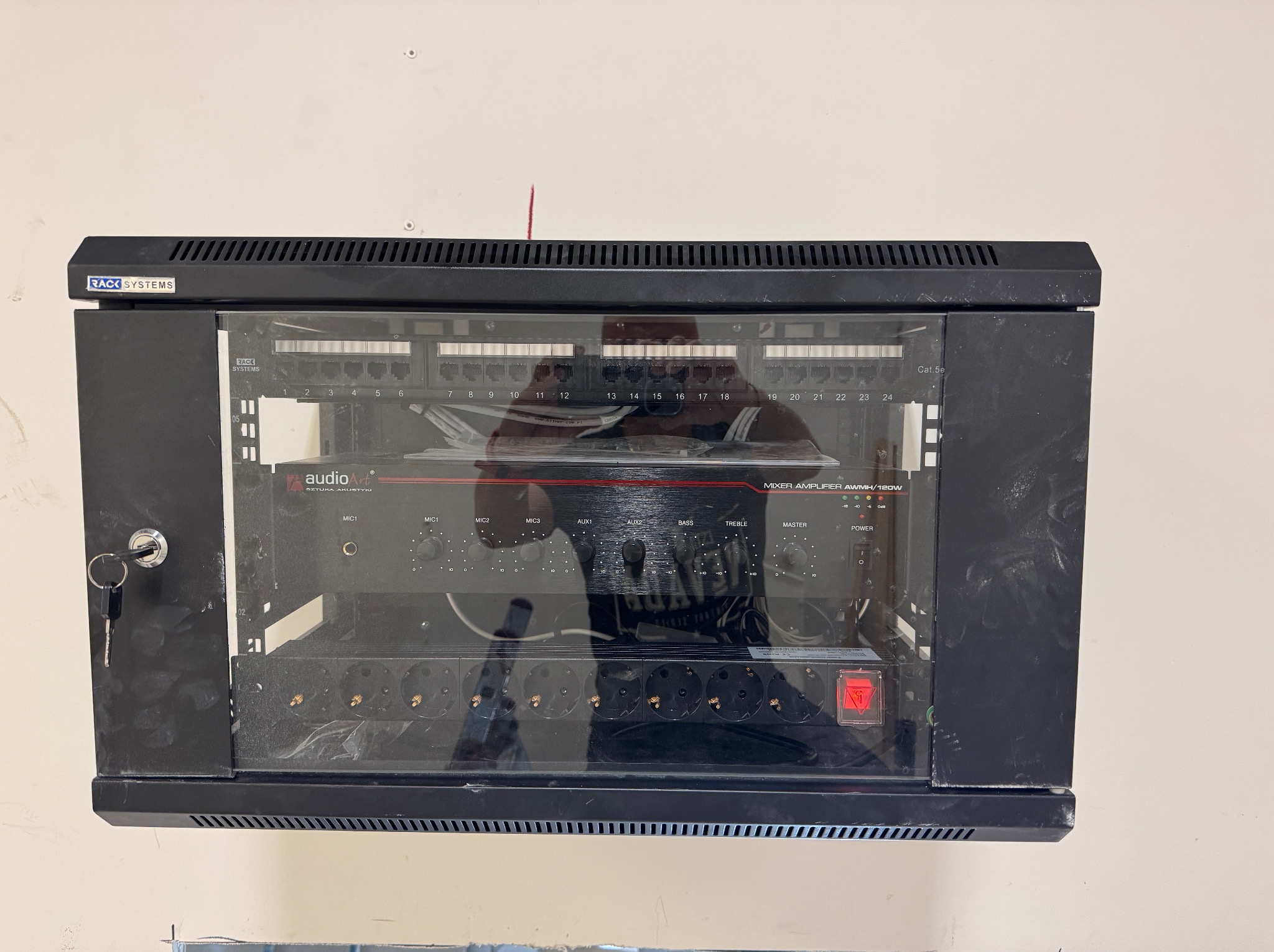 Czarna szafa rack z panelem krosowniczym, wzmacniaczem audio i listwą zasilającą, zamontowana na ścianie. Widoczny klucz w zamku szafy.