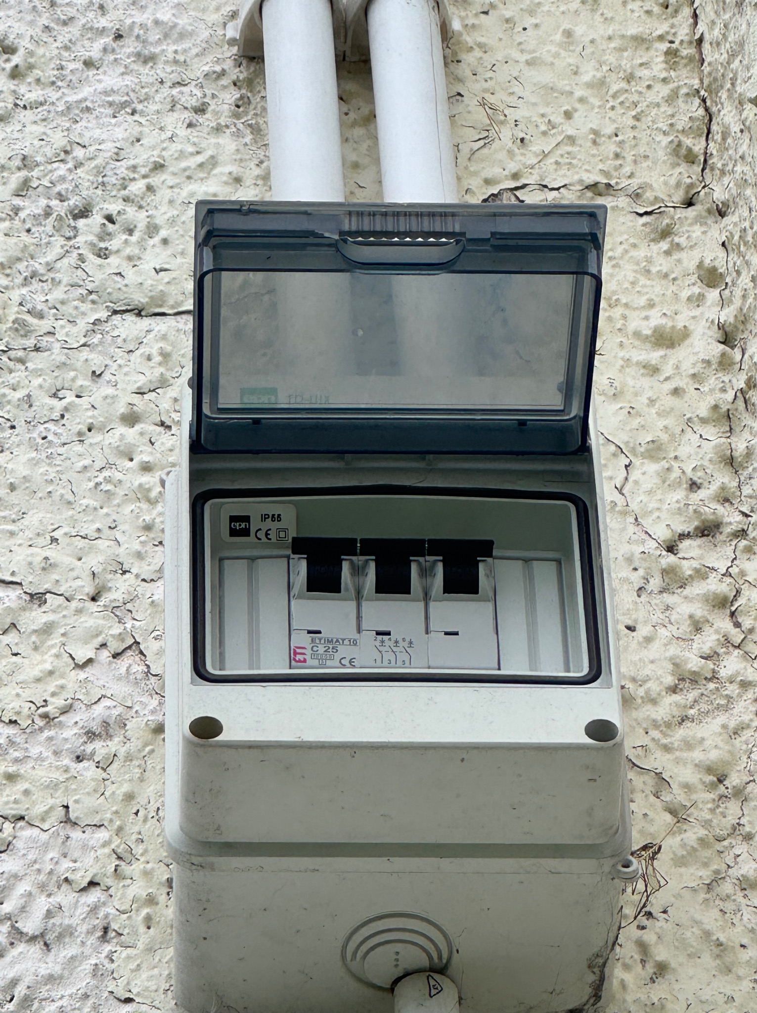 Szara skrzynka elektryczna z otwartą klapką na chropowatej, łuszczącej się ścianie. Dwie białe rury prowadzą do góry. Widoczne bezpieczniki wewnątrz.