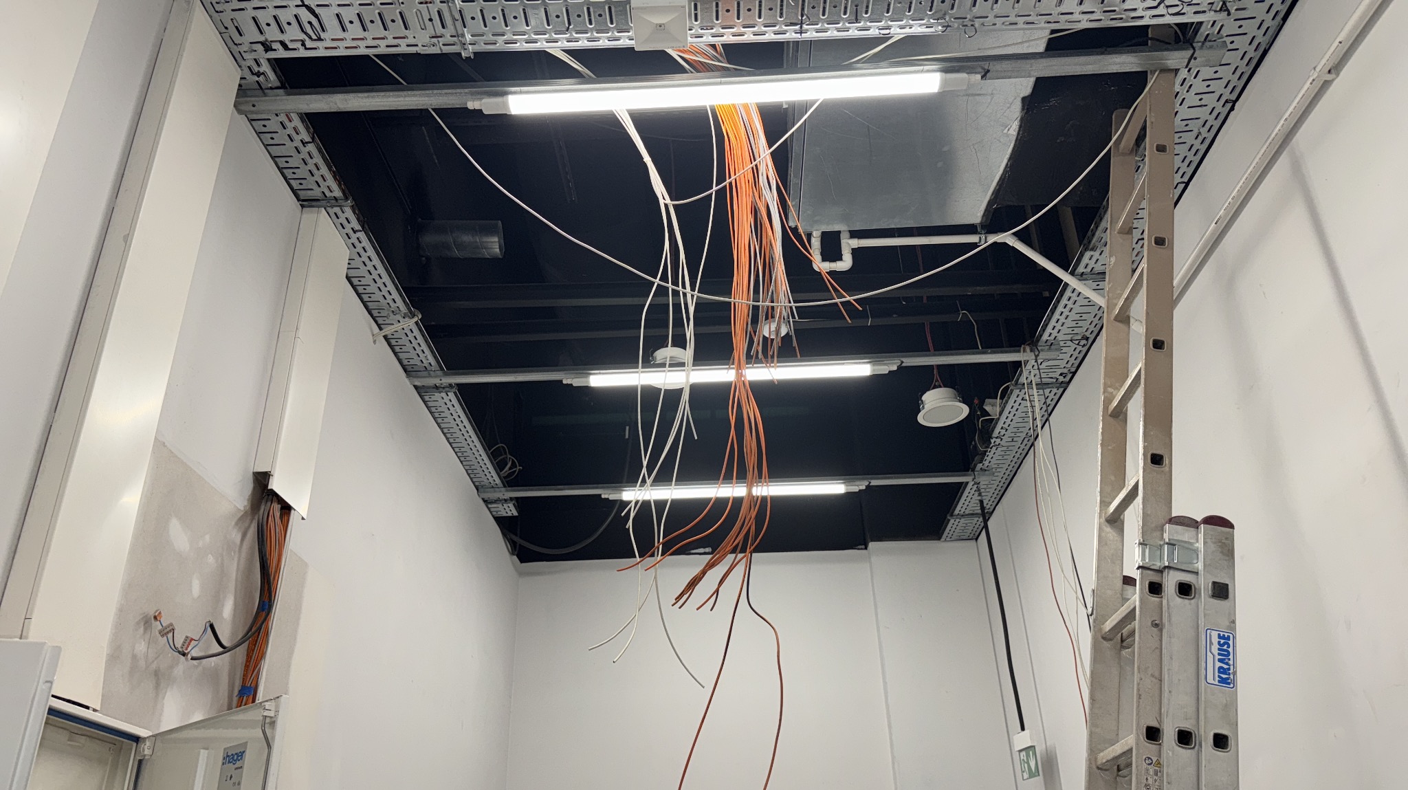 Montaż kabli w przestrzeni pod stropem podwieszanym z użyciem drabiny. Widoczne koryta kablowe i fragment szafy elektrycznej przy ścianie. Etap prac instalacyjnych.