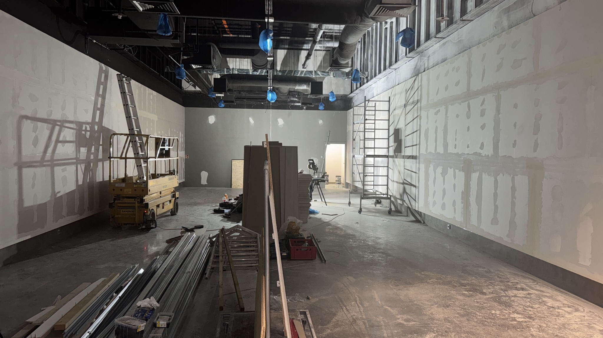 Wnętrze w trakcie wykańczania: ściany z płyt gipsowych z widocznymi łączeniami, podnośnik nożycowy, drabiny i materiały budowlane. Sufit z instalacjami.