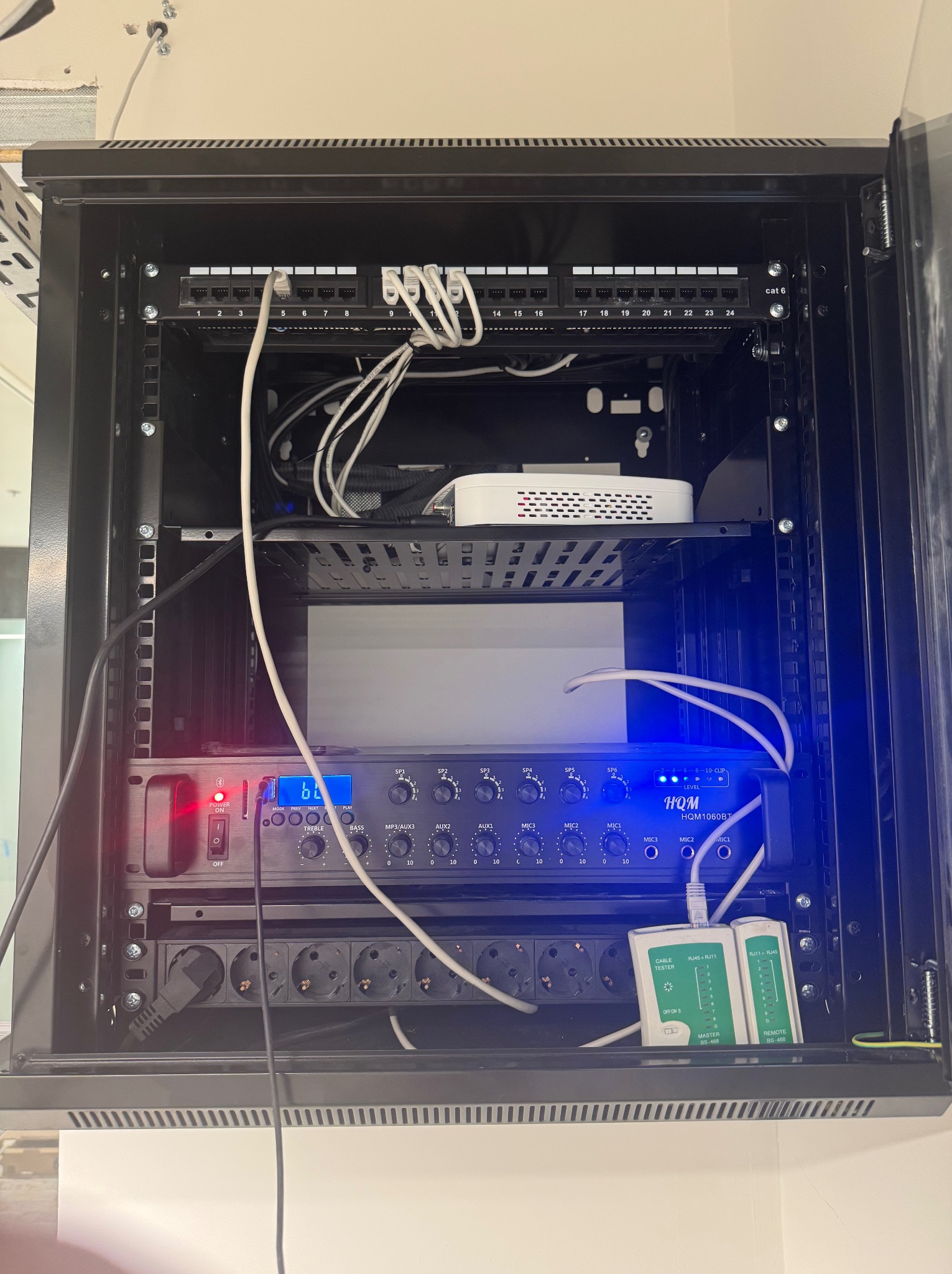 Szafa rack z patch panelem, routerem Wi-Fi, wzmacniaczem audio HQM i testerem kabli. Widoczne gniazda zasilające i kable. Oświetlenie LED podkreśla elementy.