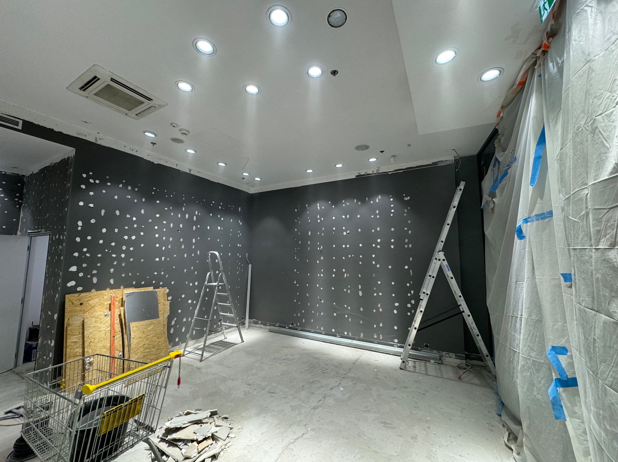 Wnętrze w trakcie prac wykończeniowych: szpachlowane ściany, widoczne drabiny, wózek z narzędziami, oświetlenie sufitowe. Prace przygotowawcze do malowania.