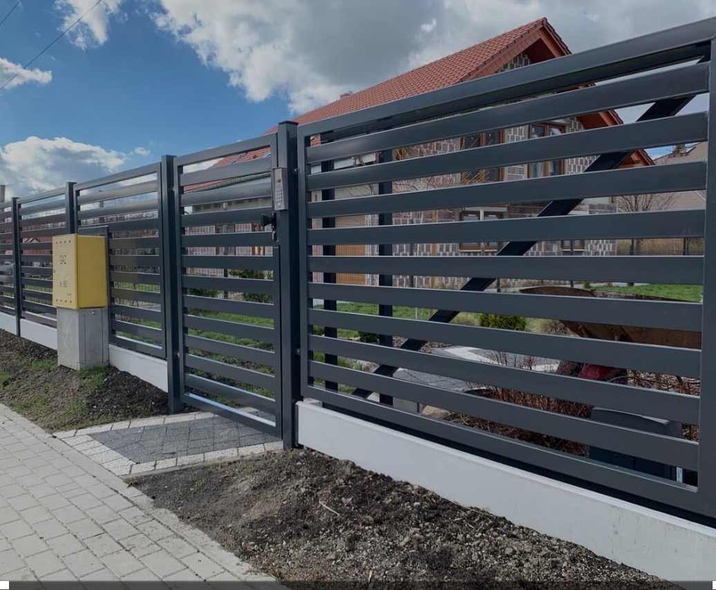 Nowoczesne, szare ogrodzenie panelowe z furtką i domofonem, widok z zewnątrz. Żółta skrzynka gazowa, kostka brukowa. Dom w tle.