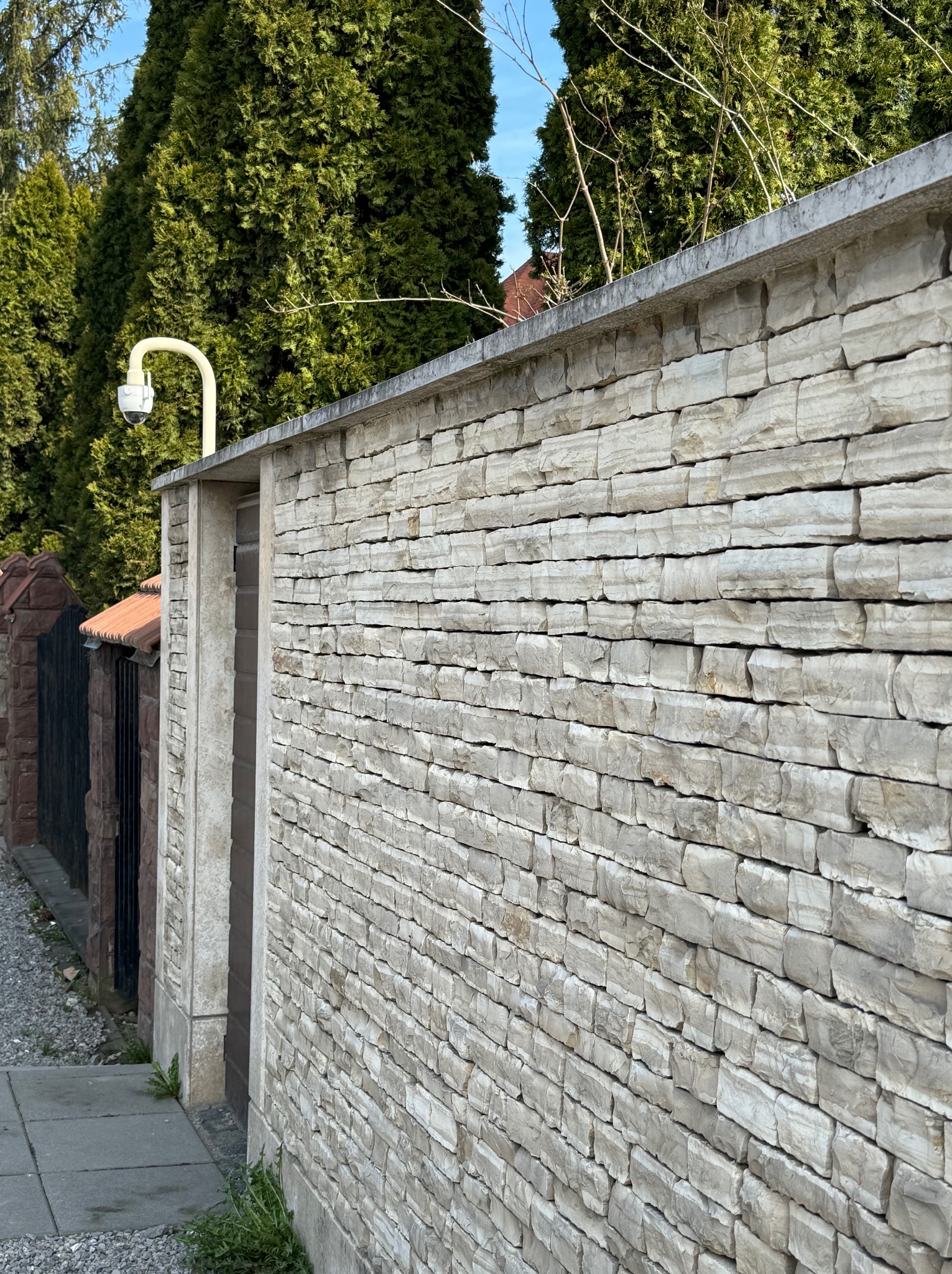Kamera monitoringu zamontowana na murze z kamienia naturalnego, obok furtki. W tle widoczne drzewa iglaste i fragment metalowego ogrodzenia.