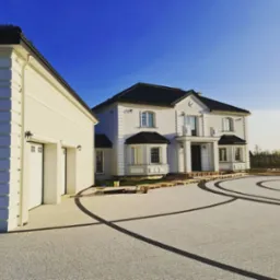 Nowoczesny dom murowany z białymi elewacjami, czarnym dachem i ozdobnymi detalami, widok z brukowanego podjazdu z ciemnymi liniami, w słoneczny dzień.