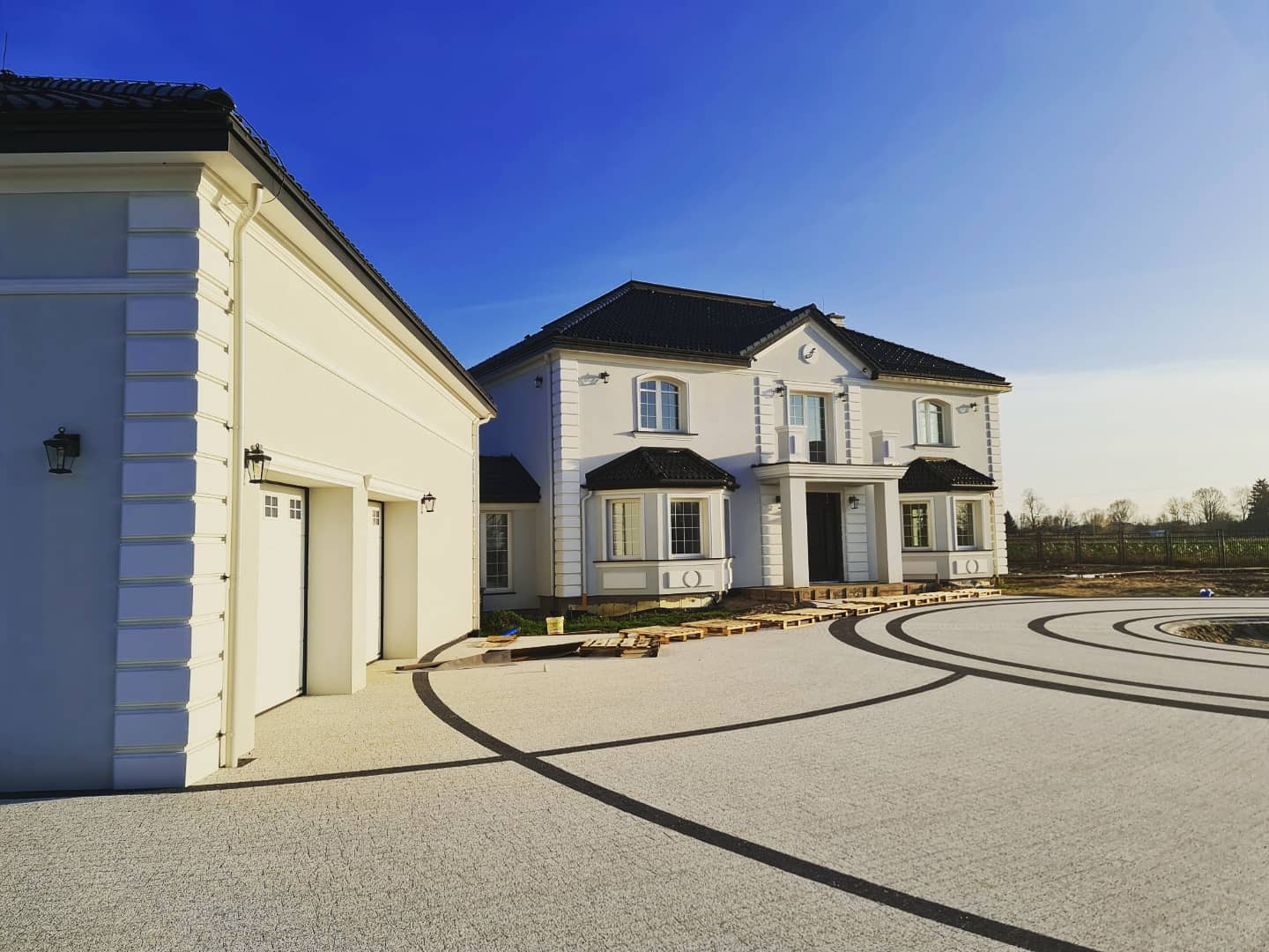 Nowoczesny dom murowany z białymi elewacjami, czarnym dachem i ozdobnymi detalami, widok z brukowanego podjazdu z ciemnymi liniami, w słoneczny dzień.