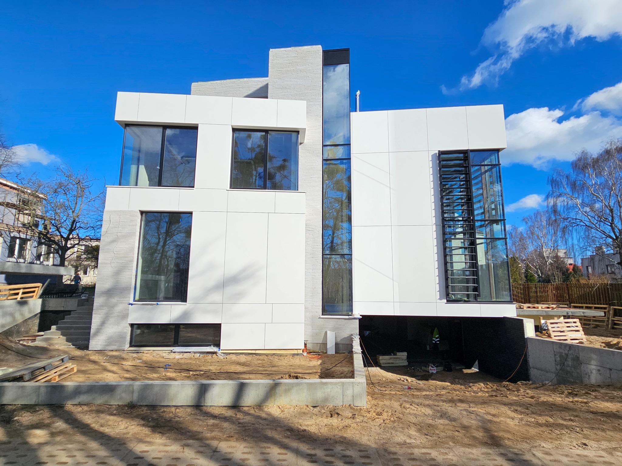 Nowoczesny dom z białymi panelami elewacyjnymi i dużymi oknami, widok z zewnątrz na plac budowy z widocznymi fundamentami i częściowo wykończonym terenem.