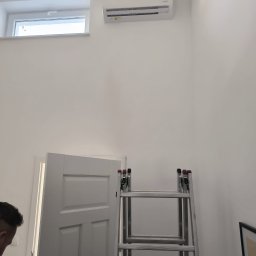 DOBRATEMPERATURA.COM SPÓŁKA Z OGRANICZONĄ ODPOWIEDZIALNOŚCIĄ - Biały klimatyzator ścienny zamontowany wysoko nad drzwiami i oknem, z rozstawioną drabiną aluminiową w pomieszczeniu o białych ścianach, widoczny fragment sylwetki osoby
