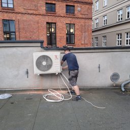 DOBRATEMPERATURA.COM SPÓŁKA Z OGRANICZONĄ ODPOWIEDZIALNOŚCIĄ - Instalator podłącza jednostkę zewnętrzną klimatyzacji Rotenso na dachu budynku, widoczne kable i elementy montażowe.