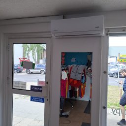 DOBRATEMPERATURA.COM SPÓŁKA Z OGRANICZONĄ ODPOWIEDZIALNOŚCIĄ - Biały klimatyzator zamontowany nad drzwiami do sklepu odzieżowego, widok z zewnątrz, z odbiciem wnętrza sklepu w szybie.