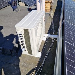 DOBRATEMPERATURA.COM SPÓŁKA Z OGRANICZONĄ ODPOWIEDZIALNOŚCIĄ - Biała jednostka zewnętrzna klimatyzacji zamontowana na płaskim dachu obok paneli słonecznych, widoczne kable i cień osoby.