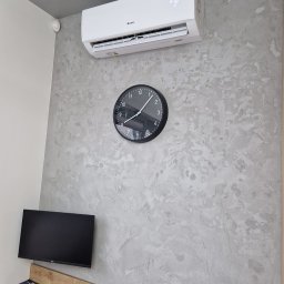 DOBRATEMPERATURA.COM SPÓŁKA Z OGRANICZONĄ ODPOWIEDZIALNOŚCIĄ - Biała jednostka klimatyzacyjna marki Gree zamontowana na dekoracyjnej, szarej ścianie obok czarnego zegara ściennego. Pod spodem widoczny biurkowy monitor Dell i klawiatura.