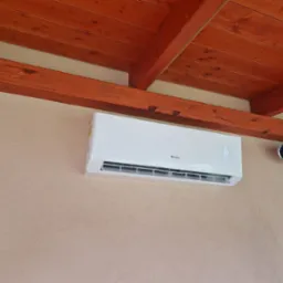 Biały klimatyzator Gree zamontowany na teksturowanej ścianie wewnątrz pomieszczenia z drewnianym sufitem i widocznym grzejnikiem zewnętrznym.