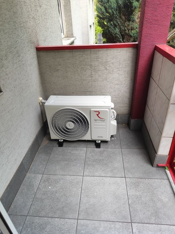 Biała jednostka zewnętrzna klimatyzatora marki Rotenso zamontowana na czarnych wspornikach na szarym balkonie z płytkami, widok z boku.