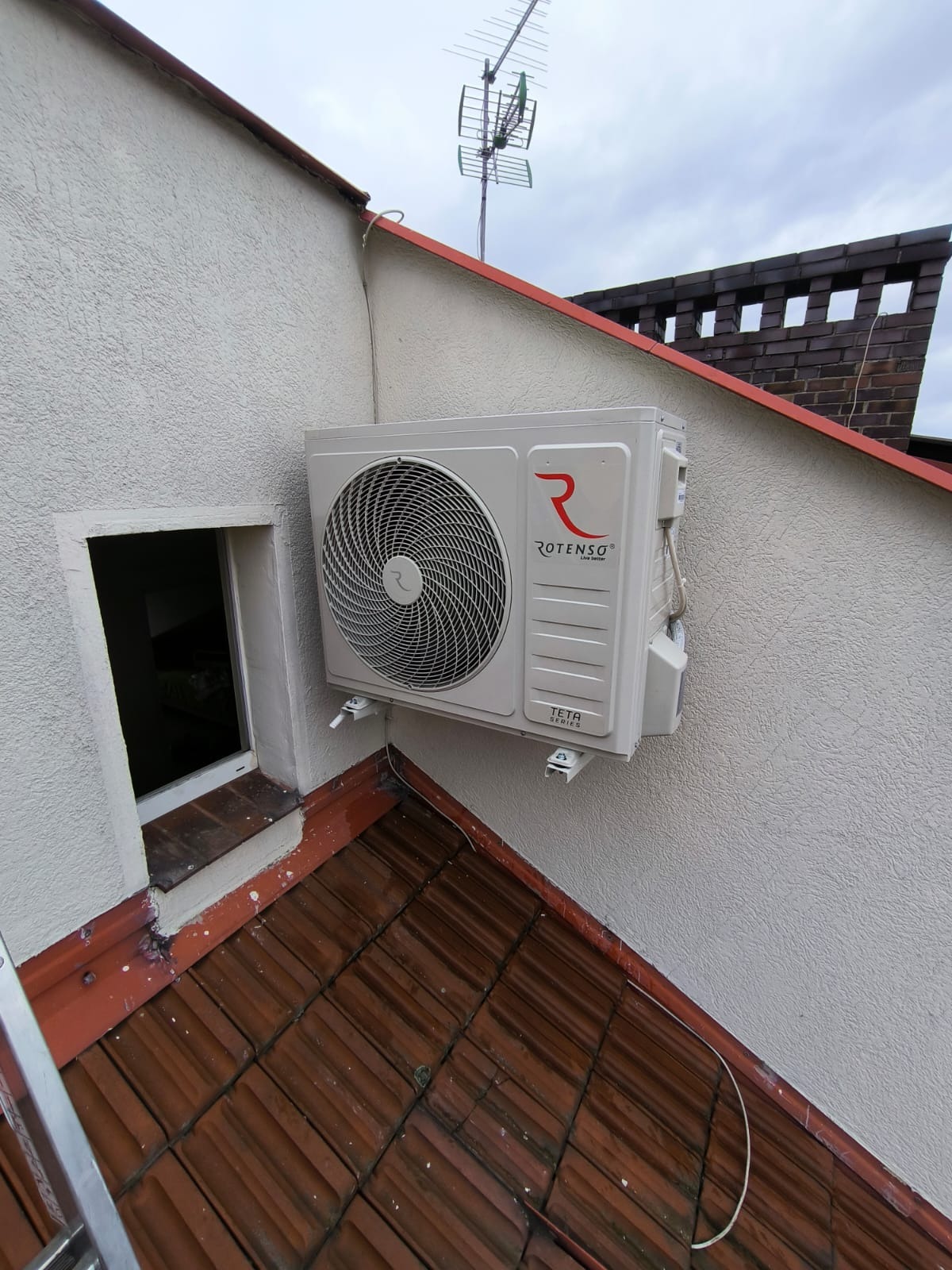 Zewnętrzna jednostka klimatyzacji Rotenso zamontowana na ścianie budynku z widoczną anteną telewizyjną na dachu i małym oknem obok.