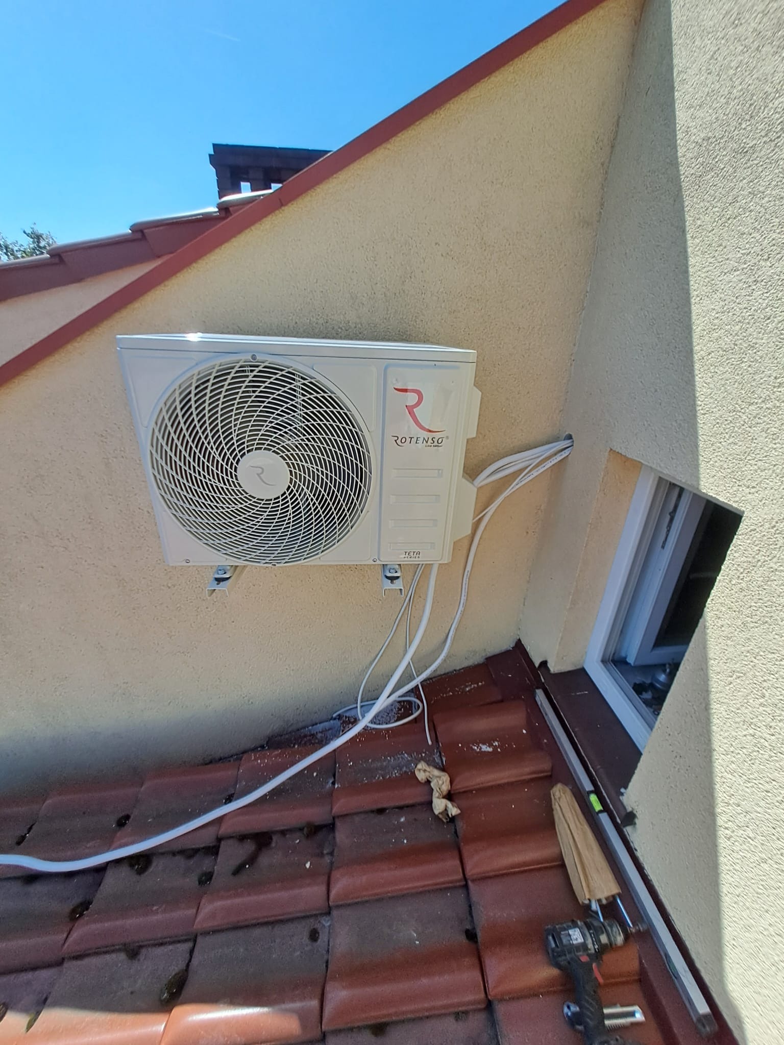 Zewnętrzna jednostka klimatyzacji Rotenso zamontowana na ścianie szczytowej budynku z widocznymi przewodami, narzędzia na dachu i okno dachowe.