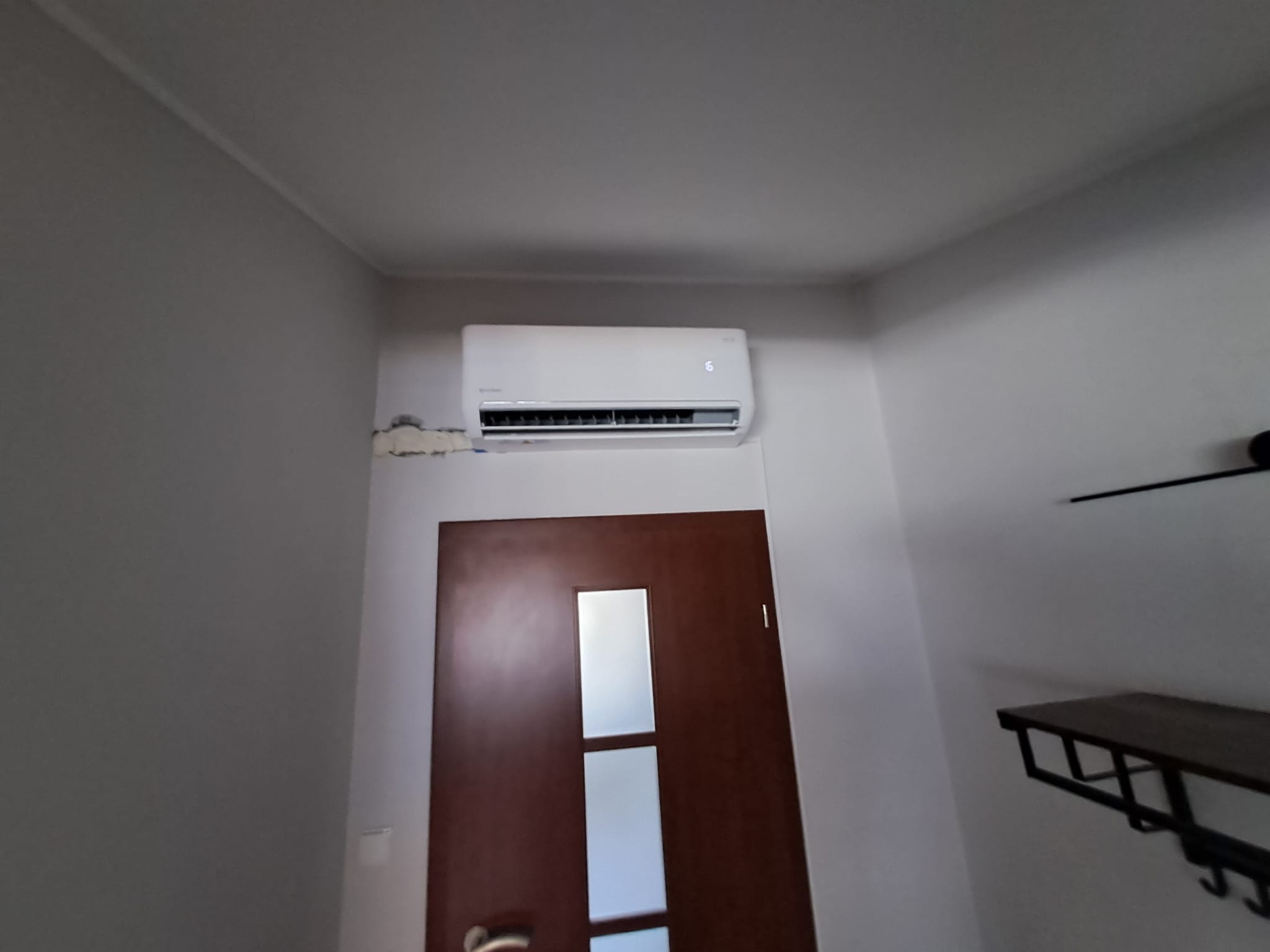Biały klimatyzator ścienny zamontowany nad brązowymi drzwiami z szybą, widoczne ślady po montażu na ścianie, ujęcie od dołu.
