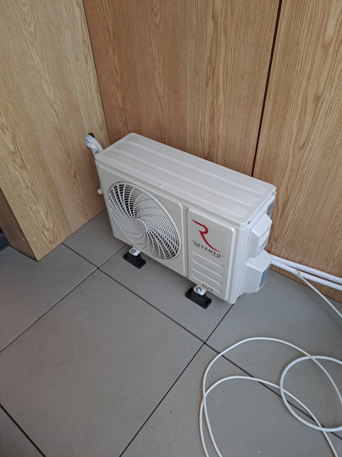 Biała jednostka wewnętrzna klimatyzacji Rotenso ELIS zamontowana na szarych płytkach podłogowych, z widocznymi przewodami chłodniczymi i odpływowymi przy drewnianej ścianie.