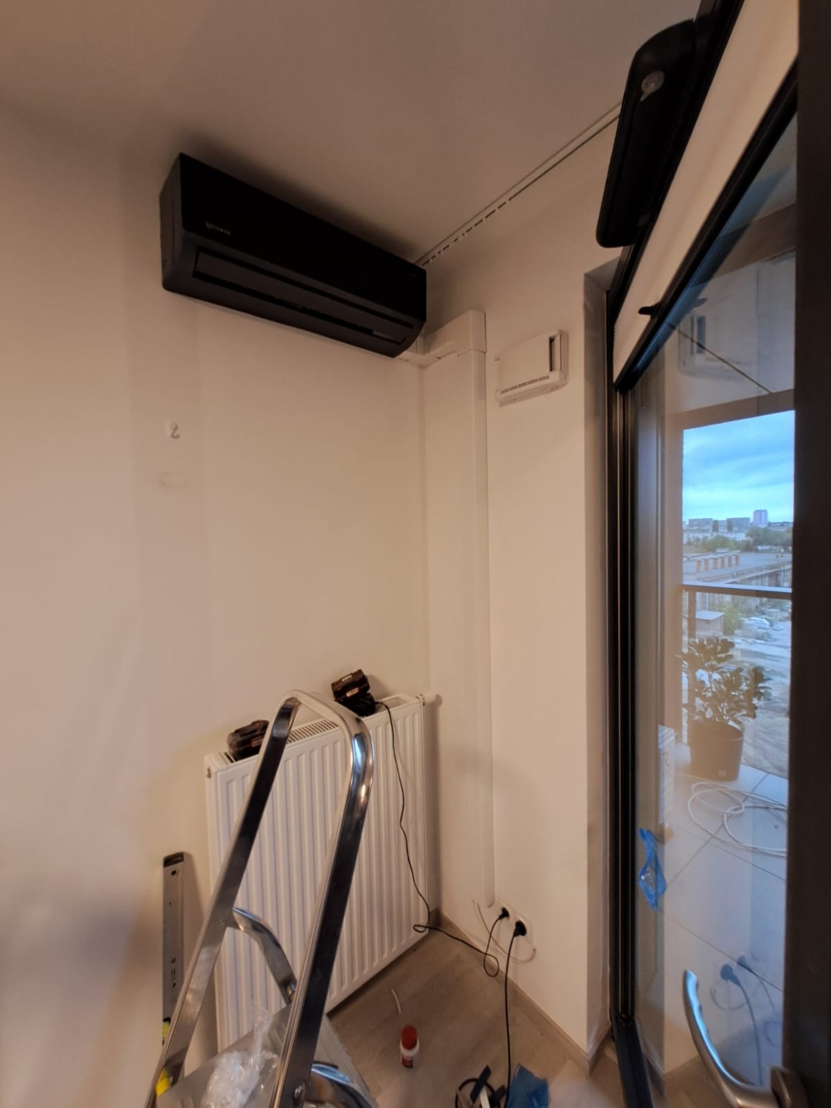 Czarny klimatyzator na ścianie w mieszkaniu, widoczne narzędzia i drabina podczas montażu, balkon z widokiem na miasto w tle.
