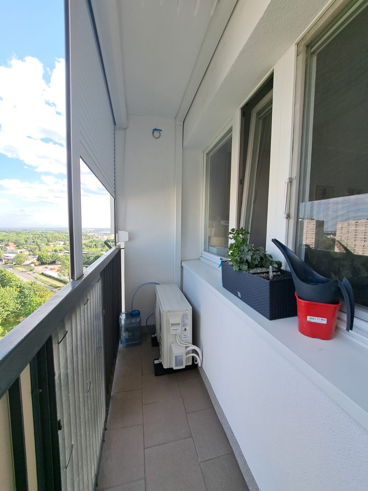 Zewnętrzna jednostka klimatyzacji na balkonie z widokiem na miasto, obok doniczka z ziołami i konewka w czerwonym wiaderku.