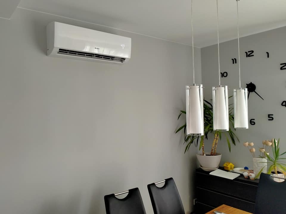 Biały klimatyzator ścienny zamontowany w szarym pokoju, widoczne lampy wiszące, zegar ścienny i meble.