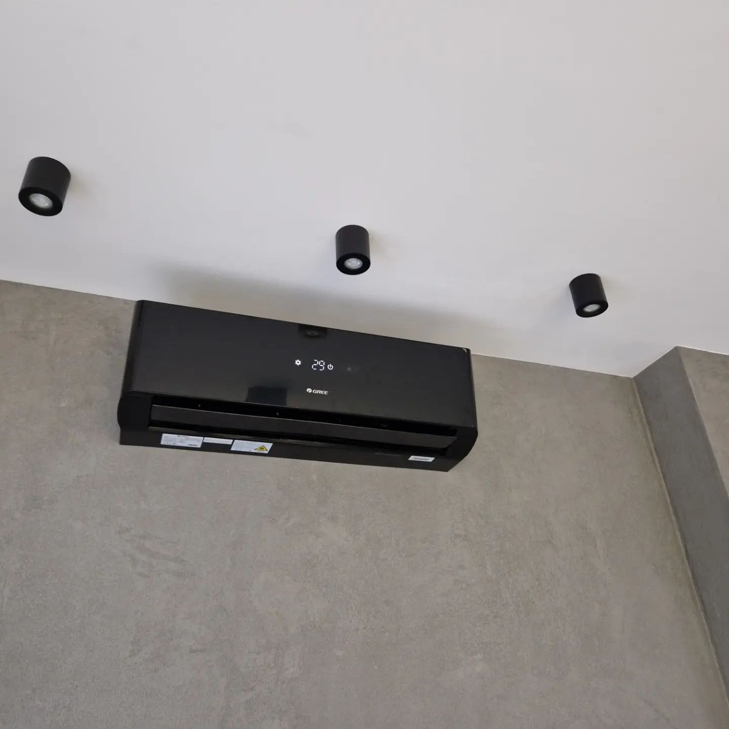 Czarny klimatyzator marki Gree zamontowany na szarej, betonowej ścianie w pomieszczeniu z trzema czarnymi lampami sufitowymi.