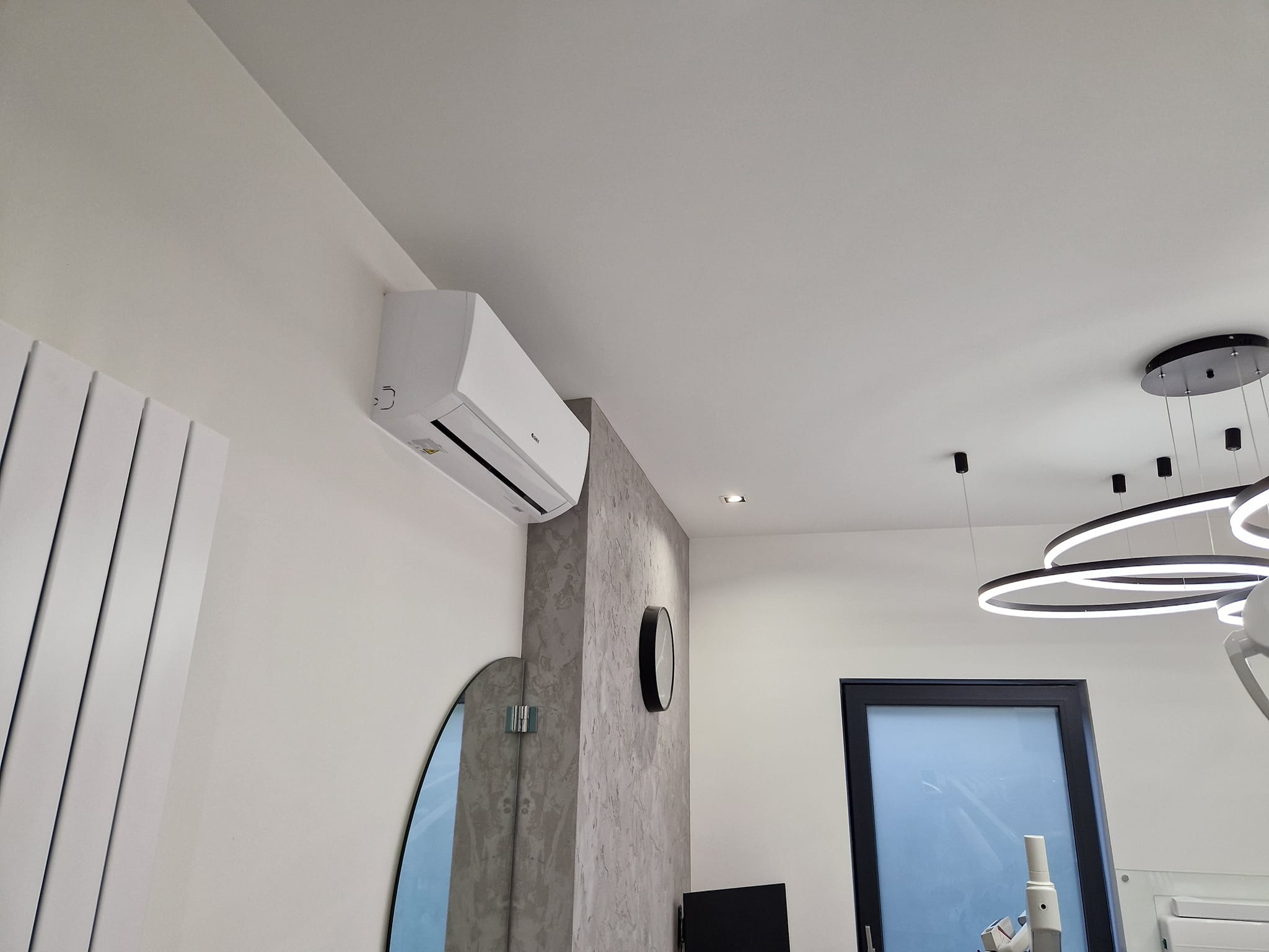 Biała jednostka klimatyzacyjna zamontowana na ścianie w nowoczesnym wnętrzu z dekoracyjnym grzejnikiem, okrągłym lustrem i designerską lampą sufitową.